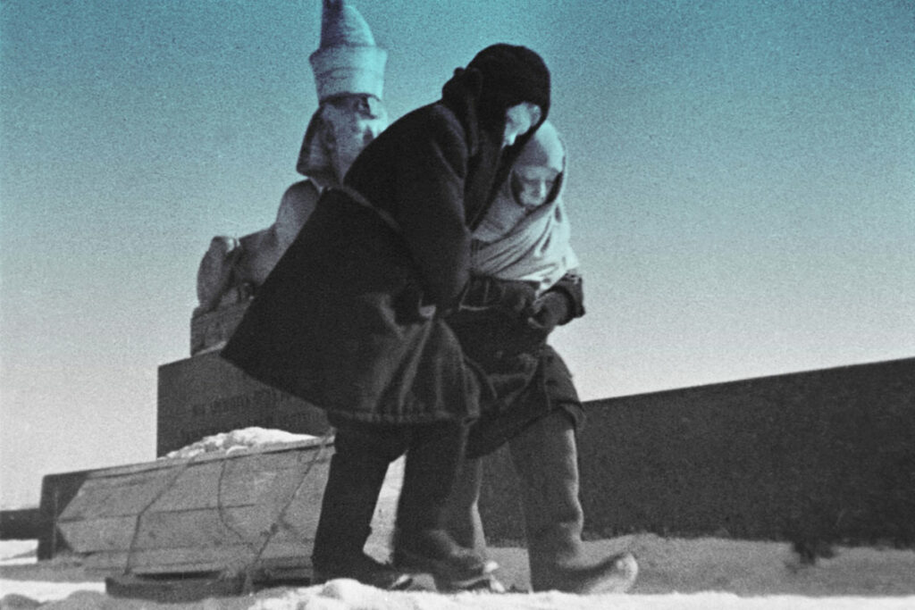 Во время блокады Ленинграда двое детей в тёплой зимней одежде везут за собой гроб по улице. На заднем плане виден памятник