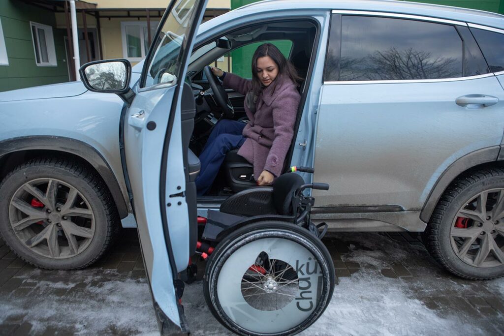 Ирина сидя в машине складывает инвалидную коляску