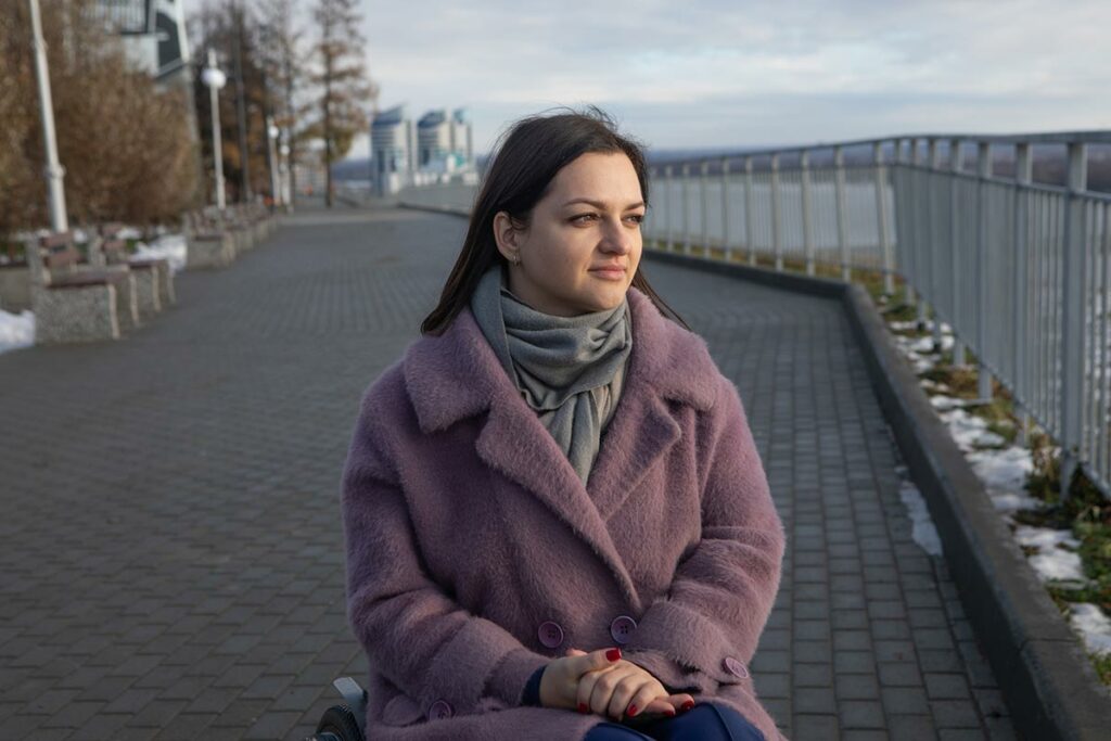 Молодая женщина в сиреневом пальто в инвалидной коляске на набережной