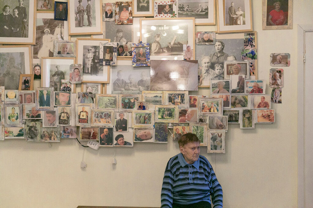 Пожилая женщина на фоне стены с фотографиями, которая в пансионате называется «стена вечной жизни»