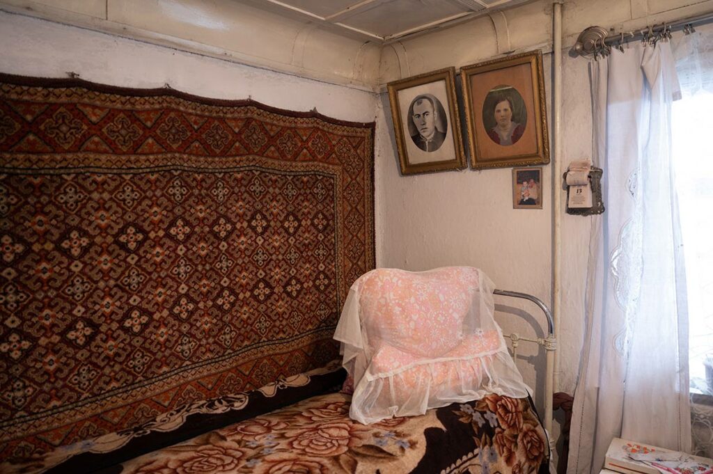 Угол в доме бабы Ани: застеленная кровать, подушки с накидкой, ковер, фотографии на стене 