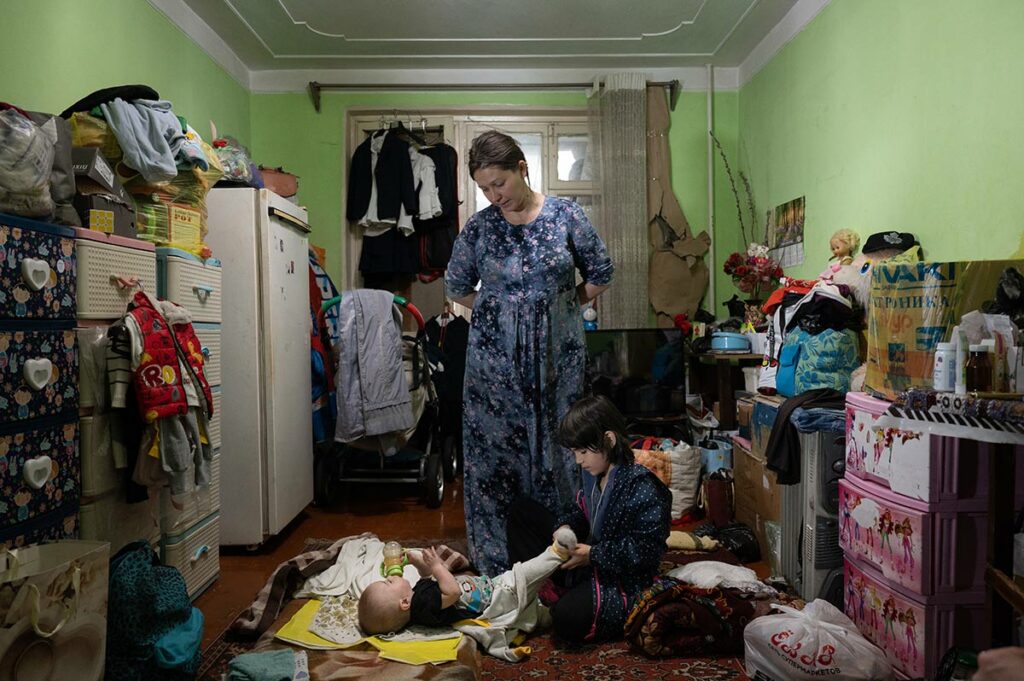 Комната, заставленная вещами по стенам. В центре стоит женщина, на полу на ковре лежит ребенок, девочка, сидящая рядом с ребенком переодевает его