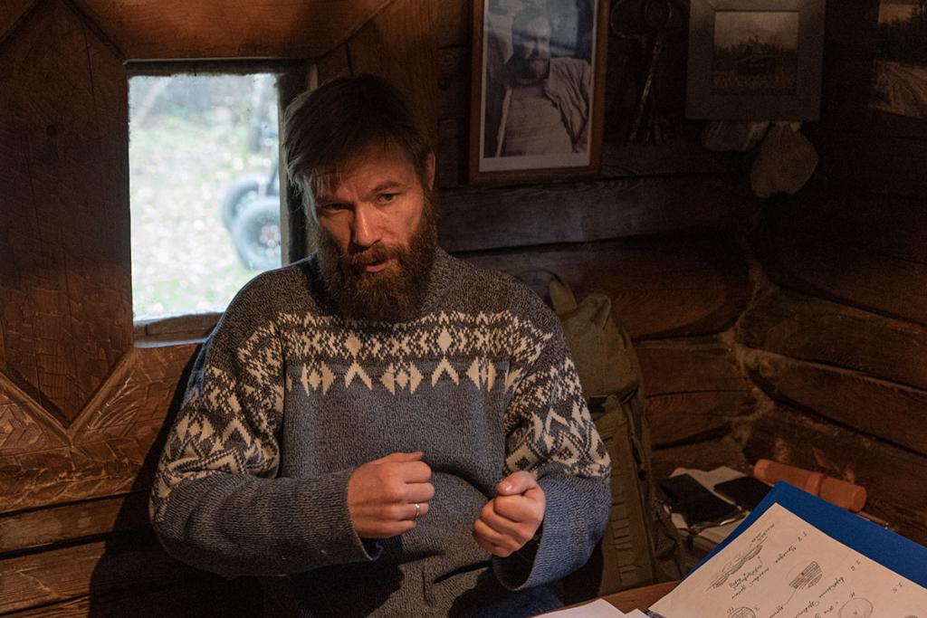Руслан Беляков, руководитель музея живой архаики «Новое Старое», в маленькой административной избушке рассказывает теорию ученикам плотницких курсов. На стене на заднем плане фото Дмитрия Александровича Соколова