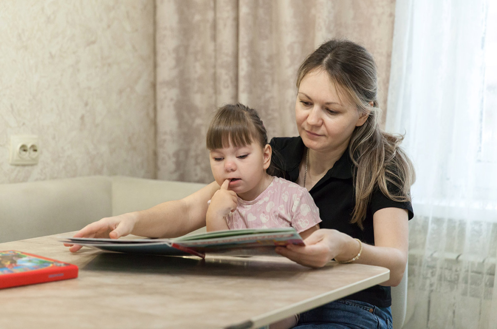 Маленькая девочка сидит за столом на коленях у мамы и смотрит в книжку 