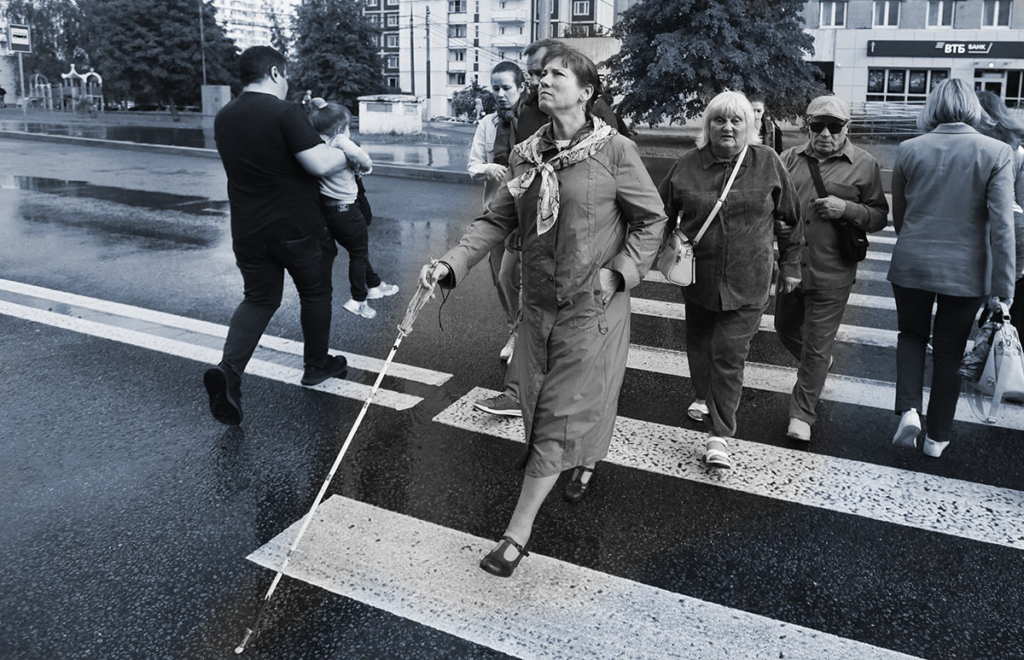 Черно-белая фотография. На первом плане слепая женщина с белой тростью