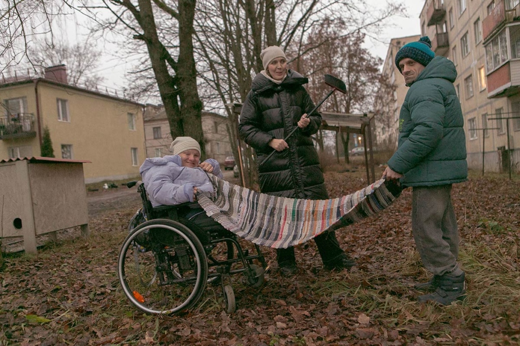 Социальный работник и двое подопечных на улице, выбивают коврик. Подопечная молодая женщина на инвалидной коляске 