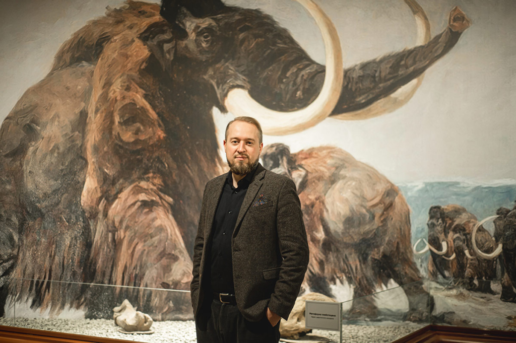 Портрет мужчины на фоне картины, изображающей мамонта