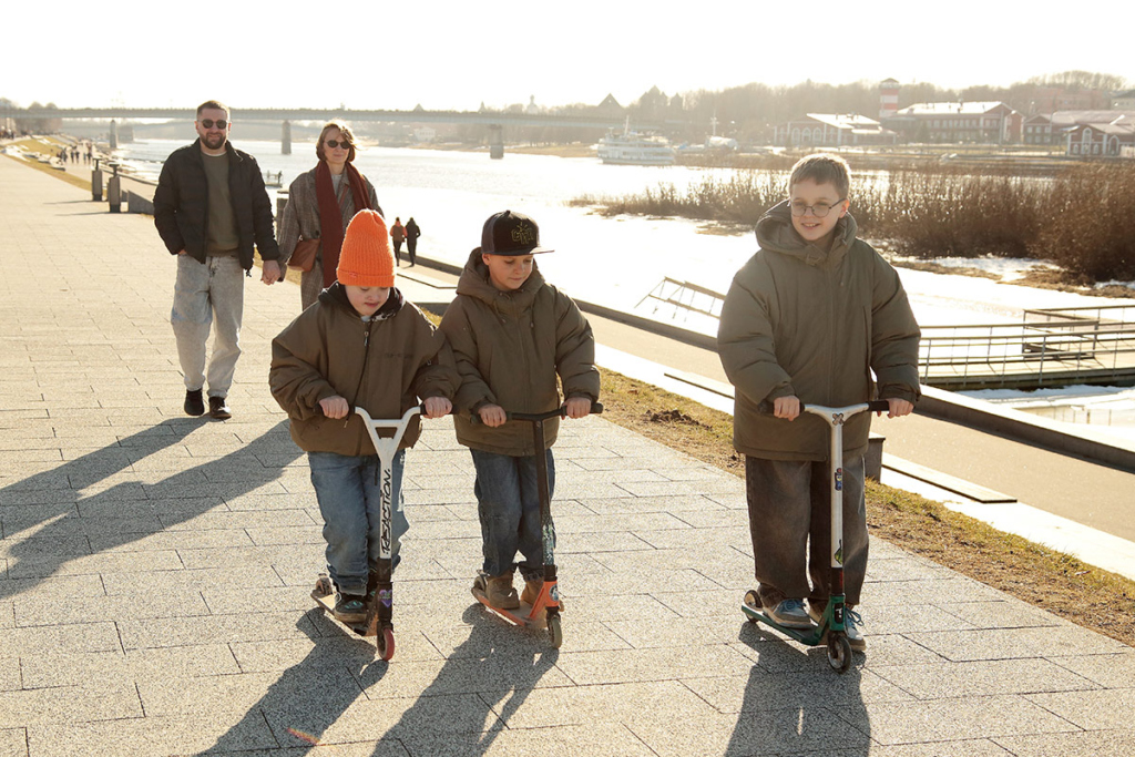 На первом плане - три мальчика едут на самокатах, за ними идут папа и мама 