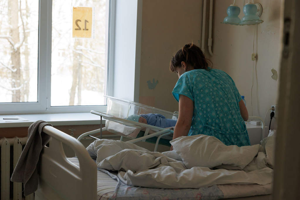 С новорождённым ребенком - 38-летняя Екатерина, третий сын Даниил. Женщина в палате роддома склонилась над кювезом младенца