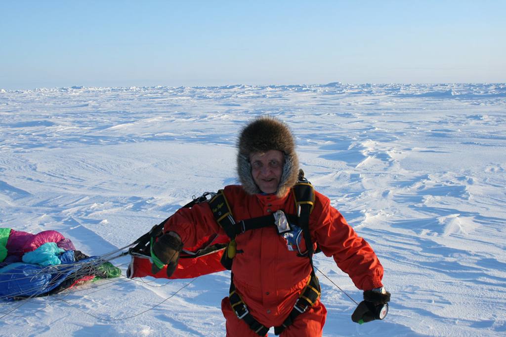 Мужчина в красном комбинезоне, в меховой шапке, за спиной сдувшийся парашют. После прыжка с парашютом. Прыжок на Северный полюс. 2012 год
