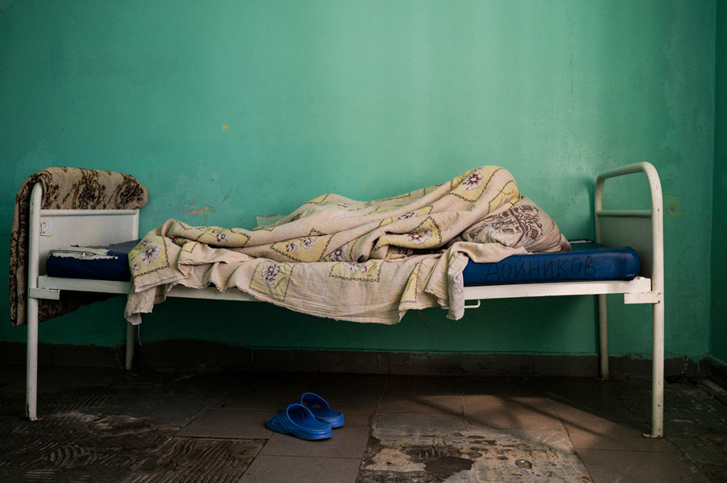 Человек лежит на кровати в ПНИ. На кровати кроме одеяла и рваного матраса ничего нет