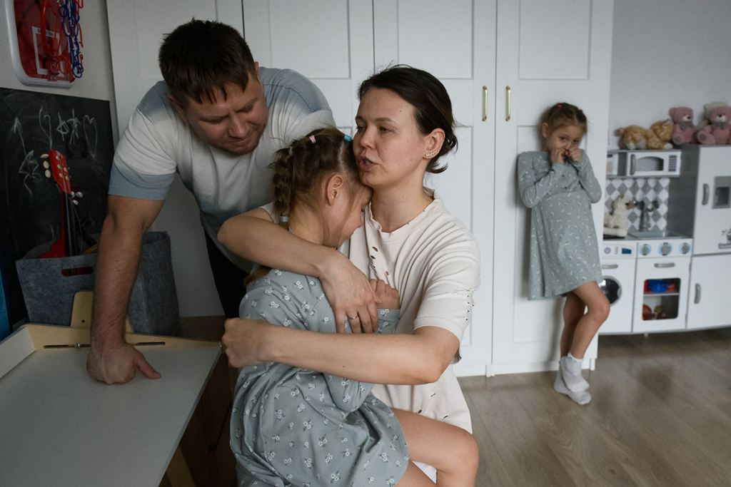 Одна дочка у мамы на руках, вторая дочка стоит в стороне у шкафа, папа рядом с мамой и дочкой