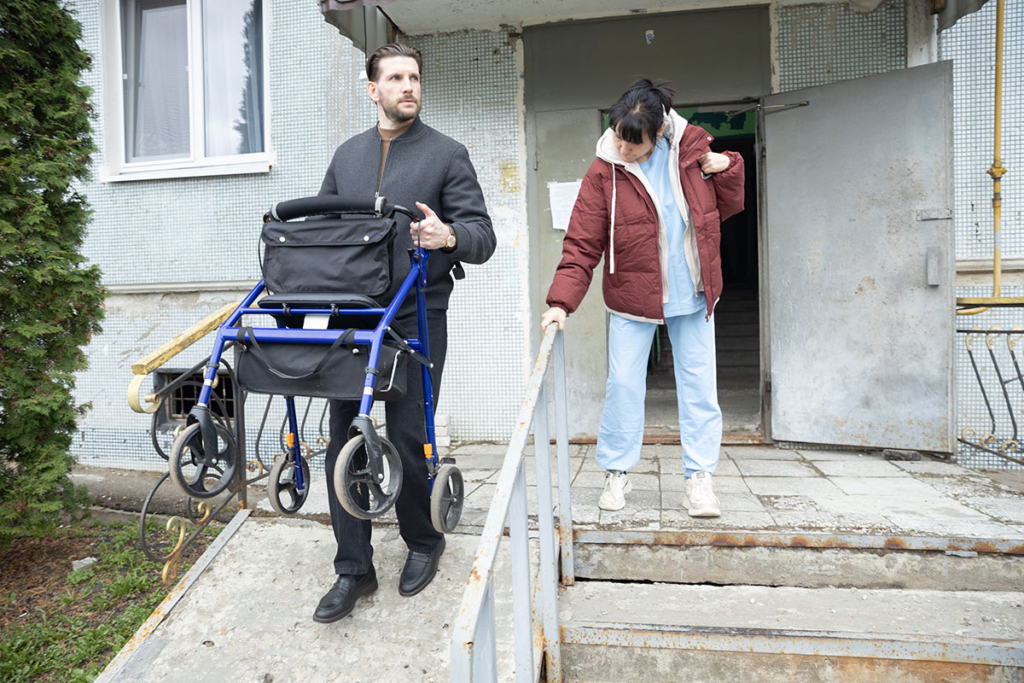 Молодой мужчина выносит ходунки из подъезда, взрослая женщина спускается по ступенькам 